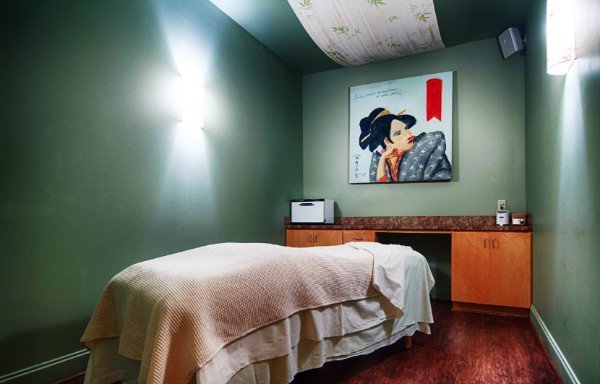 A massage room in Earthling Day Spa, Charleston. A massage table covered with sheets and a blanket in the center of the room with a large painting of an Asian girl hanging on the wall. Dim lighting and sage color walls add to the aesthetic.